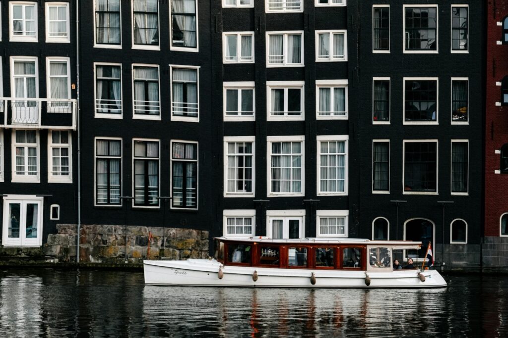 Obraz przedstawia klasyczne kamienne budynki w Amsterdamie z characteristicznym niskim, szerokim kształtem, z czarną fasadą i białymi oknami. Pod budynkami na kanale plynie biały, luksusowy STATEK. Na Pinterest można tworzyć tablice inspiracyjne dla polskich użytkowników, wykorzystując obrazy takich uroczych, historycznych miejsc. Można skupić się na kameralnych wycieczkach po miastach, podawać informacje o architekturze i kulturze, a także inspirować do podróży. W ten sposób można pomóc polskim użytkownikom znaleźć nowe pomysły dla swoich podróży, projektów domowych lub hobby związanych z turystyką.