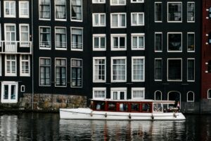 Obraz przedstawia klasyczne kamienne budynki w Amsterdamie z characteristicznym niskim, szerokim kształtem, z czarną fasadą i białymi oknami. Pod budynkami na kanale plynie biały, luksusowy STATEK. Na Pinterest można tworzyć tablice inspiracyjne dla polskich użytkowników, wykorzystując obrazy takich uroczych, historycznych miejsc. Można skupić się na kameralnych wycieczkach po miastach, podawać informacje o architekturze i kulturze, a także inspirować do podróży. W ten sposób można pomóc polskim użytkownikom znaleźć nowe pomysły dla swoich podróży, projektów domowych lub hobby związanych z turystyką.